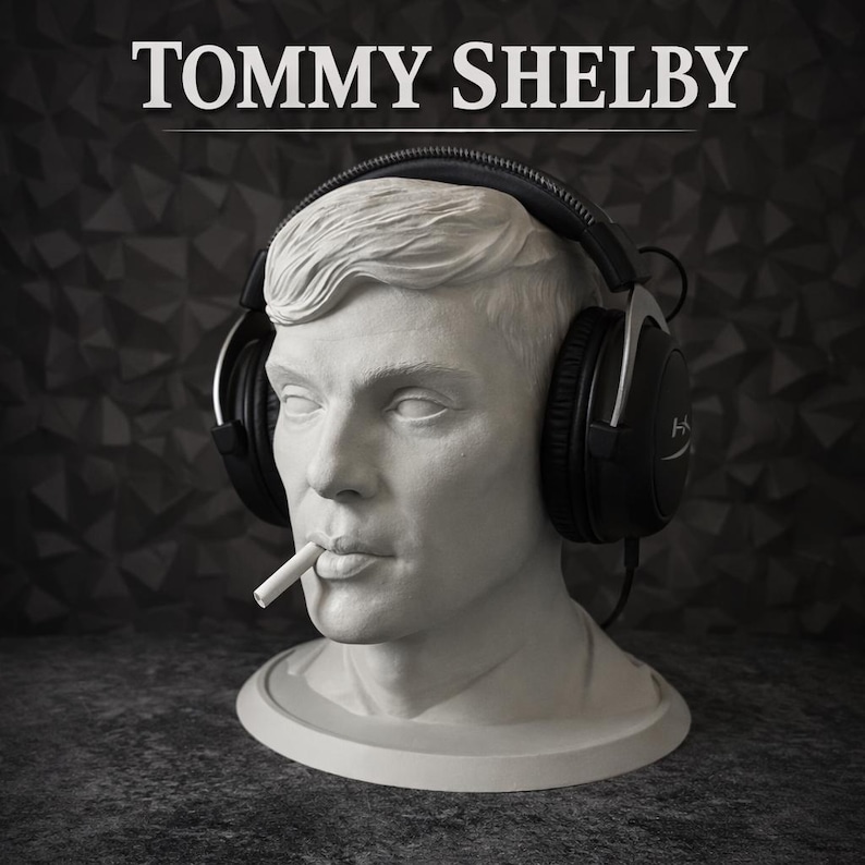 May include: A white bust of a man with a cigarette in his mouth, wearing black over-ear headphones. The text TOMMY SHELBY is printed in white above the head. The sculpture features detailed facial features and hair, resting on a circular base.