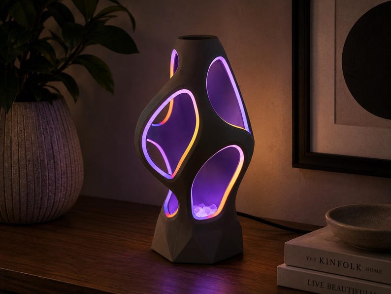 K&ouml;nnte beinhalten: Eine skulpturale Tischleuchte mit einer matten, grauen, organisch geformten Au&szlig;enh&uuml;lle mit mehreren Aussparungen. Die Innenkanten der Aussparungen sind mit lebhaftem violettem und orangefarbenem LED-Licht beleuchtet, das auf kleine klare Steine strahlt.