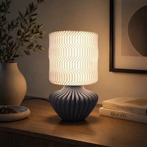 K&ouml;nnte beinhalten: Eine Tischleuchte mit einem zylindrischen wei&szlig;en Lampenschirm mit vertikalem Zickzackmuster und einem matten grauen Fu&szlig; mit tiefen, abgerundeten vertikalen Falten. Die Lampe strahlt ein warmes, sanftes Licht aus, das die texturierte Oberfl&auml;che beleuchtet.