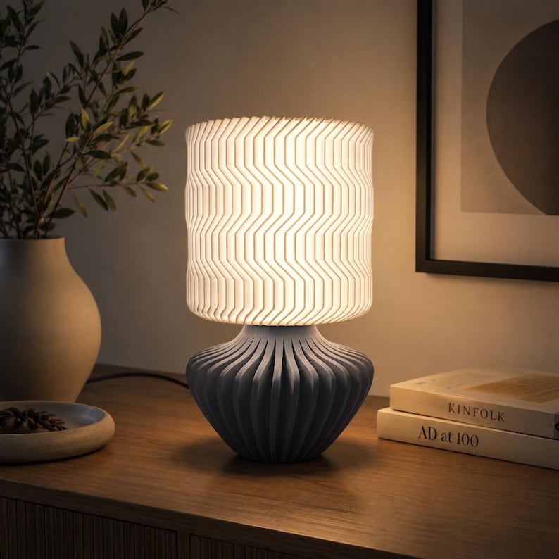 K&ouml;nnte beinhalten: Eine Tischleuchte mit einem zylindrischen wei&szlig;en Lampenschirm mit vertikalem Zickzackmuster und einem matten grauen Fu&szlig; mit tiefen, abgerundeten vertikalen Falten. Die Lampe strahlt ein warmes, sanftes Licht aus, das die texturierte Oberfl&auml;che beleuchtet.