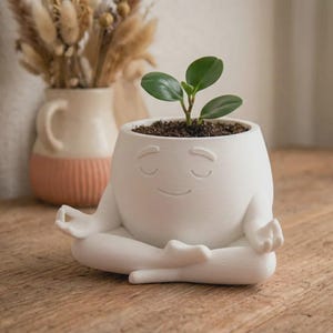 May include: A white ceramic planter shaped like a figure sitting in a cross-legged meditation pose with eyes closed and a gentle smile. The planter holds a small green plant with rounded leaves, and the surface has a smooth, matte finish.