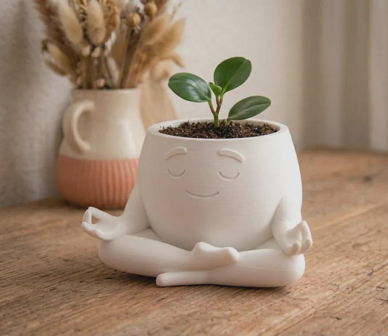 May include: A white ceramic planter shaped like a figure sitting in a cross-legged meditation pose with eyes closed and a gentle smile. The planter holds a small green plant with rounded leaves, and the surface has a smooth, matte finish.