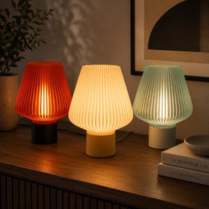 K&ouml;nnte beinhalten: Drei Tischlampen mit gerippten, glockenf&ouml;rmigen Schirmen in Rot, Gelb und Hellgr&uuml;n. Jede Lampe hat einen massiven zylindrischen Sockel in Schwarz, Gelb beziehungsweise Wei&szlig;. Die Lampen strahlen ein warmes, sanftes Licht aus und beleuchten die Umgebung.