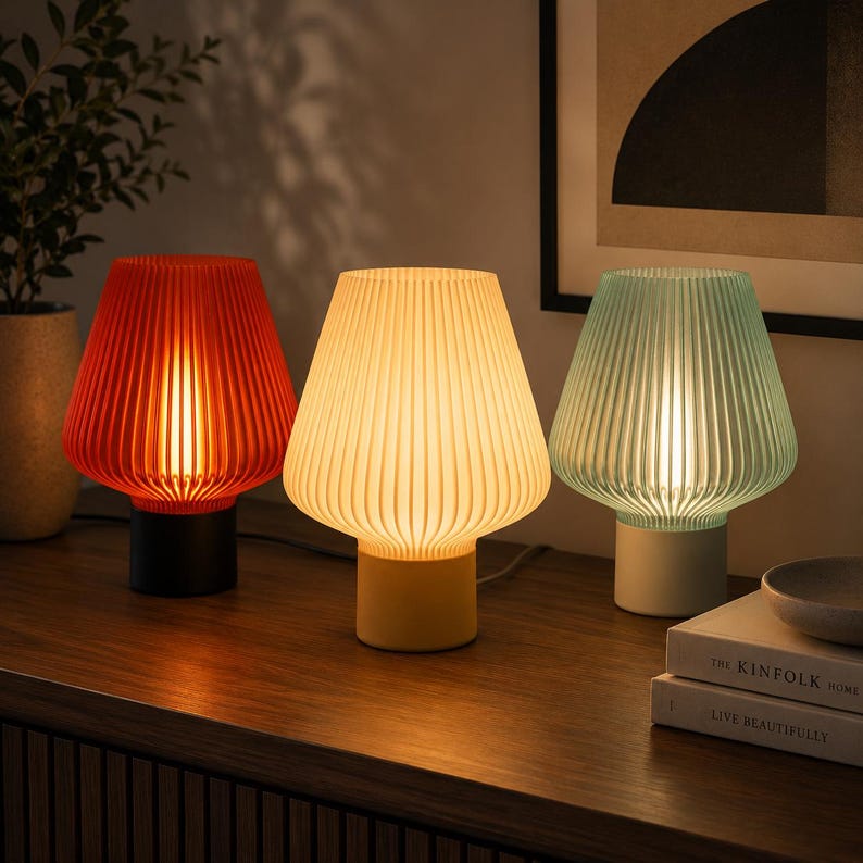 K&ouml;nnte beinhalten: Drei Tischlampen mit gerippten, glockenf&ouml;rmigen Schirmen in Rot, Gelb und Hellgr&uuml;n. Jede Lampe hat einen massiven zylindrischen Sockel in Schwarz, Gelb beziehungsweise Wei&szlig;. Die Lampen strahlen ein warmes, sanftes Licht aus und beleuchten die Umgebung.