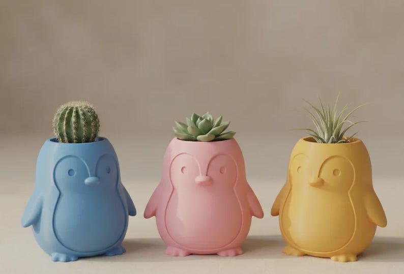 May include: Three penguin-shaped planters in blue, pink, and yellow. The blue planter holds a round green cactus, the pink planter contains a green succulent, and the yellow planter holds a spiky green air plant. Each planter has a smooth finish.