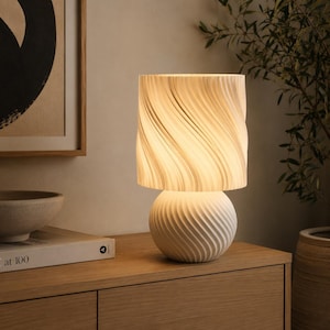 K&ouml;nnte beinhalten: Eine Tischleuchte mit einem kugelf&ouml;rmigen Fu&szlig; mit diagonal gerippter Textur und einem zylindrischen Lampenschirm mit vertikalem, wellenf&ouml;rmigem Plisseemuster. Die Lampe strahlt ein warmes, sanftes Licht aus, das die wei&szlig;e Oberfl&auml;che beleuchtet.