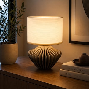 K&ouml;nnte beinhalten: Eine Tischleuchte mit einem gefalteten, beigen, f&auml;cherf&ouml;rmigen Fu&szlig; und einem zylindrischen, cremewei&szlig;en Stoffschirm. Die Lampe strahlt ein warmes, sanftes Licht aus, das die Umgebung erhellt. Der Fu&szlig; hat vertikale Falten mit Textur.