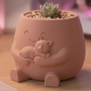 May include: A peach-colored ceramic planter shaped like a rounded figure with a smiling face, holding a small sculpted cat in its arms. The planter contains a light green succulent plant nestled in small, light-colored decorative pebbles.