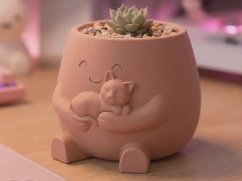 May include: A peach-colored ceramic planter shaped like a rounded figure with a smiling face, holding a small sculpted cat in its arms. The planter contains a light green succulent plant nestled in small, light-colored decorative pebbles.