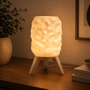 K&ouml;nnte beinhalten: Eine wei&szlig;e Tischleuchte mit einem strukturierten, geometrischen und facettierten Lampenschirm, der ein warmes, sanftes Licht ausstrahlt. Die Leuchte steht auf drei stabilen, angewinkelten wei&szlig;en Beinen, die der zylindrischen Form Halt geben.