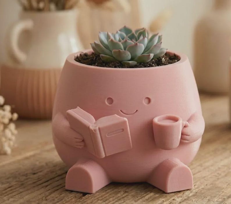 May include: A matte pink planter shaped like a seated character holding an open book in one hand and a small mug in the other. The planter features a simple embossed face with two circular eyes and a curved smile, containing a green succulent plant.