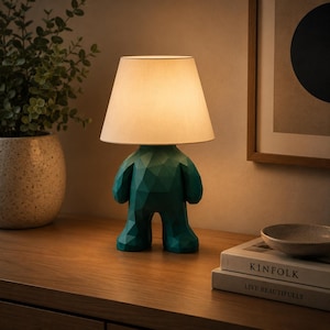 K&ouml;nnte beinhalten: Eine Tischleuchte mit einem dunkel-t&uuml;rkisfarbenen, geometrischen, menschen&auml;hnlichen Sockel und einem einfachen, cremewei&szlig;en, konischen Stoffschirm. Die Lampe strahlt ein warmes, sanftes Licht aus und beleuchtet die Umgebung gezielt.