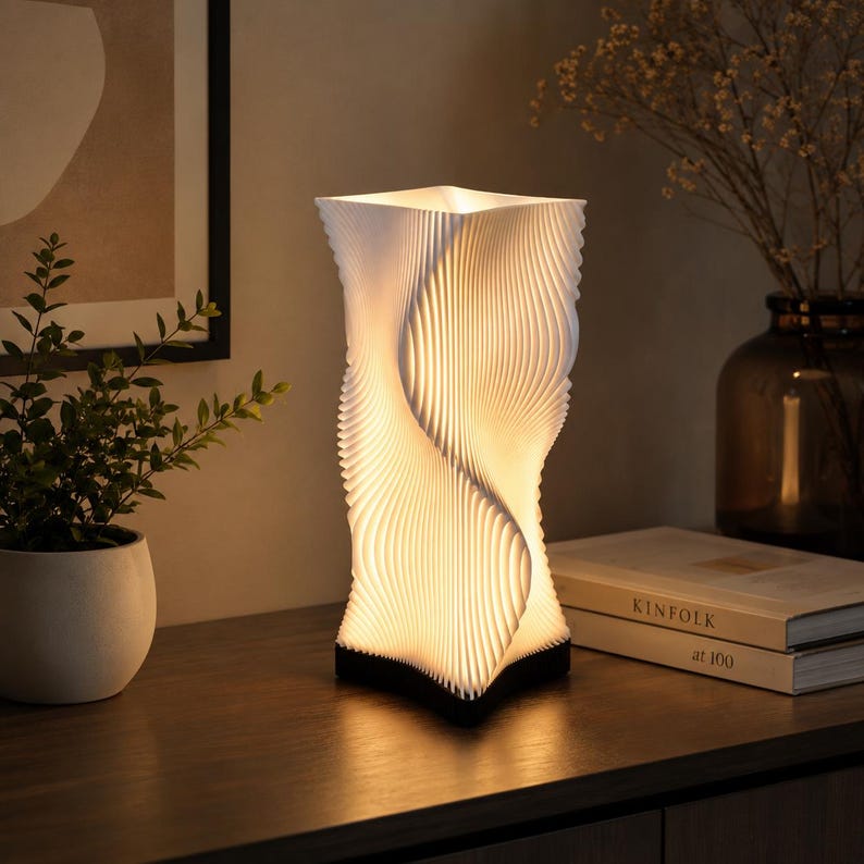 K&ouml;nnte beinhalten: Eine wei&szlig;e Tischleuchte mit einer hohen, rechteckigen und vertikal gerippten Struktur, die ein gedrehtes, wellenartiges Muster erzeugt. Die Lampe strahlt ein warmes, sanftes Licht von innen aus, das die geschichtete Textur des Designs betont.