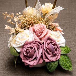 Peut inclure: Un arrangement floral composé de roses mauve foncé et crème, agrémenté de petits boutons beiges, de feuillage séché et de feuilles vertes. La composition présente un mélange de pétales à la texture douce et d'éléments botaniques aériens.