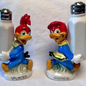 May include: A pair of vintage ceramic salt and pepper shakers featuring the cartoon character Woody Woodpecker. Each shaker is a white rectangular bottle held by a Woody Woodpecker figurine. One is labeled "Splinter" and the other "Knothhead".