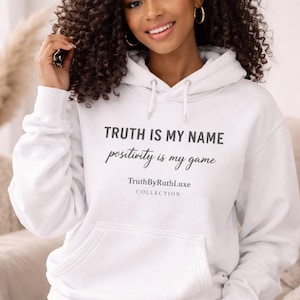 May include: A white hoodie with a front pocket and drawstring. The text "TRUTH IS MY NAME positivity is my game" is printed on the front. The hoodie is from the TruthByRuthLuxe COLLECTION.
