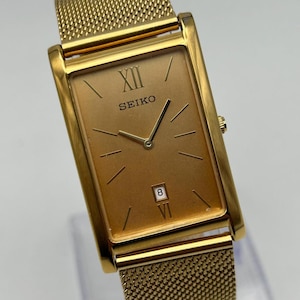 May include: A gold-toned Seiko watch with a rectangular face and mesh band. The watch face displays Roman numerals, the brand name, and the date. The watch has a gold-colored case and band.
