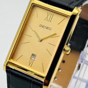 Könnte beinhalten: Goldfarbene Seiko-Uhr mit rechteckigem Zifferblatt und schwarzem Lederarmband. Das Zifferblatt hat römische Ziffern, Strichmarkierungen und ein Datumsfenster. Die Uhr hat eine goldfarbene Lünette und ein strukturiertes schwarzes Lederarmband.