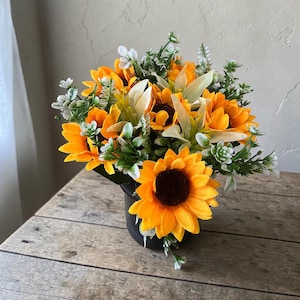 May include: A vibrant floral arrangement featuring bright yellow sunflowers with dark brown centers, accented by white and green foliage. The flowers are arranged in a black pot, creating a cheerful and natural aesthetic. The arrangement sits on a wooden surface.