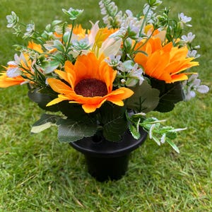 Könnte beinhalten: Ein lebendiges Blumenarrangement mit leuchtend orangefarbenen Sonnenblumen mit dunkelbraunen Zentren, akzentuiert durch weiße und grüne Blätter. Die Blumen sind in einer schwarzen Vase angeordnet, vor einem Hintergrund aus grünem Gras.