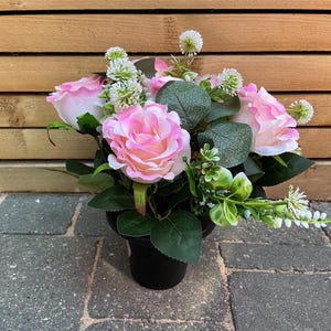 May include: An artificial floral arrangement featuring light pink and white roses, small white spherical flower clusters, and green eucalyptus leaves. The flowers are contained within a simple black pot, creating a compact and balanced display.