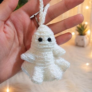 May include: A small white crocheted ghost ornament featuring a rounded head, two black plastic bead eyes, and a ruffled, flared base. The ghost is crafted from white yarn and includes a matching crocheted loop at the top for hanging purposes.