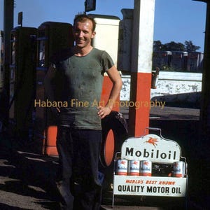 Peut inclure: Photographie vintage d'un homme devant une station-service Mobil. L'enseigne indique "Mobiloil, World's Best Known, Quality Motor Oil". Plusieurs bidons d'huile sont exposés dans un présentoir métallique. L'homme porte une chemise verte et un pantalon sombre.