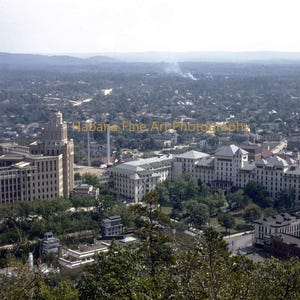 Peut inclure: Vue aérienne d'une ville avec plusieurs grands bâtiments et une haute tour. Les bâtiments sont de couleur claire et entourés d'arbres. Le ciel est brumeux, et l'ensemble de la scène est un peu délavé.
