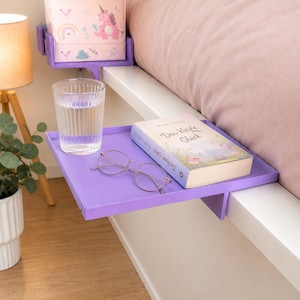 Könnte beinhalten: Ein lila Nachttischregal, das an einem weißen Bettrahmen befestigt ist und ein Glas Wasser, ein Buch und eine Brille enthält. Ein zweites Regal mit einer dekorativen Box ist im Hintergrund zu sehen. Das Buch trägt den Titel "Das Kleine Glück".