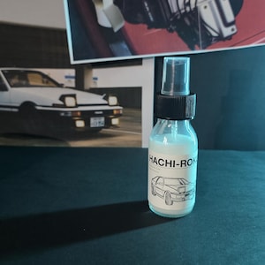 May include: A clear glass spray bottle with a black cap and the text "HACHI-ROK" in white. The bottle contains a white liquid and features a car illustration. Background includes car-related posters and a dark surface.