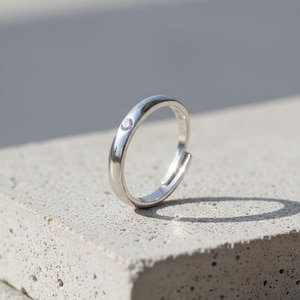 Puede incluir: Un anillo ajustable de plata con una pequeña gema transparente. El anillo está sobre una superficie gris texturizada, con una sombra detrás. El diseño del anillo es simple y elegante.