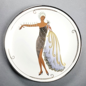 May include: A round white ceramic plate with a silver rim featuring a central illustration of a figure in a dark, textured, one-shoulder dress with a long, flowing, light blue and gold-trimmed cape, wearing a white headpiece and dark shoes.