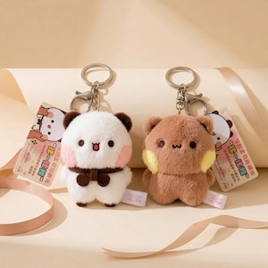 May include: Two plush animal keychains with metal rings and lobster clasps. One is white with brown ears and a brown scarf, the other is brown with yellow cheek patches. Both feature small black eyes and mouths, and include attached paper tags with text.