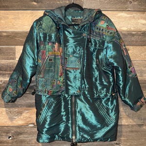 May include: Teal iridescent winter coat with a hood. The coat has a full-length zip, pockets, and decorative patches with colourful geometric designs. The coat is hanging on a black hanger against a wooden background.