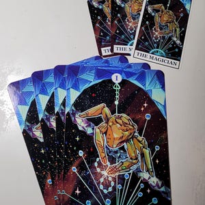 May include: A set of tarot cards featuring The Magician card, which displays a golden, mechanical-looking figure against a dark, starry space background with blue geometric patterns and the text THE MAGICIAN printed at the bottom of each card.