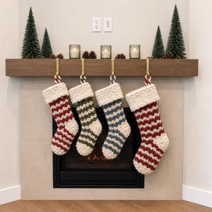 May include: Four chunky knit Christmas stockings hanging from a wooden mantelpiece. Each stocking features a thick cream-colored cuff and a striped pattern in alternating colors of red, dark green, blue, and red, all suspended by gold metal hooks.