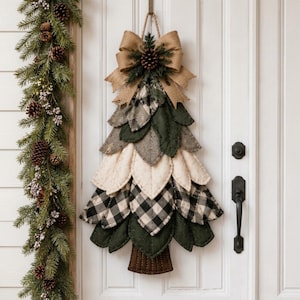 May include: A hanging wall decoration shaped like a Christmas tree, constructed from layered fabric leaves in shades of dark green, cream, and black and white buffalo plaid. It features a brown fabric trunk and a large burlap bow with a pinecone.