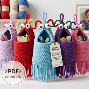 May include: Five hanging crochet organisers in purple, red, light blue, dark purple, and pink. Each features two large white eyes, colourful pipe cleaner antennae, a wide open mouth for storage, and a bottom fringe. A tag reads Cuddly Monster.