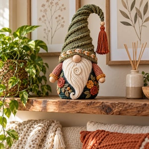 May include: A crochet gnome figurine featuring a tall, spiraled olive green hat with a rust-colored tassel, a round wooden nose, and a long cream-colored beard. The body is dark green with embroidered floral accents in rust and yellow.