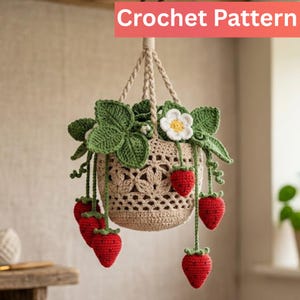May include: A hanging crochet plant basket in beige yarn with a decorative openwork pattern. The basket contains crocheted green leaves, a white flower with a yellow center, and several dangling red strawberries with small black seed details.
