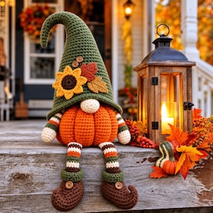 May include: A crocheted gnome figure with a tall olive green hat adorned with a yellow sunflower and two fabric leaves. The gnome has a round orange pumpkin body, striped arms and legs, and brown curled-toe shoes with small wooden buttons.
