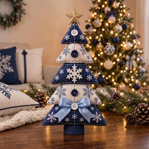 Peut inclure: Une décoration de sapin de Noël en tissu composée de cinq segments triangulaires empilés, alternant entre le bleu marine et le crème, avec des motifs de flocons blancs, des boutons blancs, un nœud en satin bleu clair et une étoile dorée.