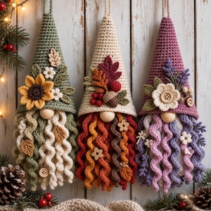 Pode incluir: Três gnomos de crochê pendurados com chapéus pontiagudos. O gnomo da esquerda é verde sálvia com um girassol, o central é creme com folhas de outono e bagas, e o da direita é malva com um botão de flor e ramos de lavanda. Todos têm barbas encaracoladas.