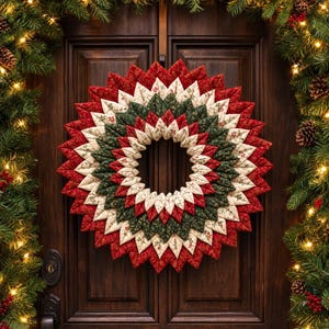 Op de afbeelding: Een ronde stoffen krans opgebouwd uit gevouwen, doorgestikte stoffen blaadjes in concentrische ringen van rood, crème en donkergroen. De stof is voorzien van kleine bloemmotieven, wat een stervormig effect creëert met een hol midden.