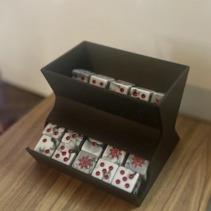 May include: A dark brown, two-tiered dice holder filled with white dice. The dice have red pips and red nautical wheel designs. The holder sits on a wooden surface.