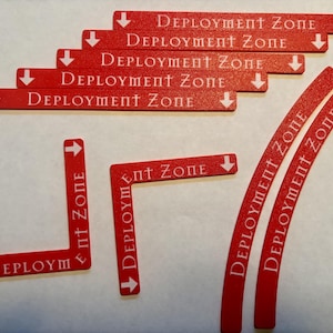 May include: Red game pieces with the text "DEPLOYMENT ZONE" in white. The pieces include straight lines, curved lines, and right-angle shapes with directional arrows. The pieces are for a board game.