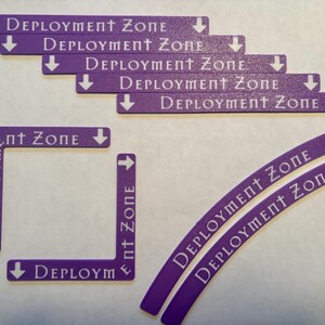May include: Purple game zone markers with white text that reads "DEPLOYMENT ZONE" and arrows. The set includes straight, curved, and corner pieces for tabletop games.