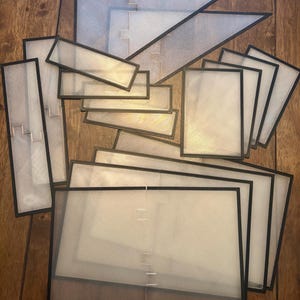 May include: A collection of clear plastic sleeves in rectangular and triangular shapes, with black borders. The sleeves are arranged on a wooden surface, some overlapping. The transparent material allows visibility of the contents within.