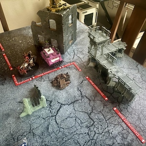 Op de afbeelding: Een tabletop-oorlogsspelscène met miniatuurstructuren en -figuren. Bevat een roze tank, een gedetailleerde figuur en verschillende gebouwmodellen. Rode markeringen met de tekst "Deployment Zone" zijn rond de scène geplaatst.