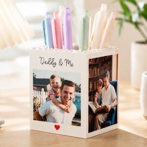 May include: A white block-shaped pen holder with a photo of a father and child, and the text "Daddy & Me". The holder contains pens in various colors. A red heart is at the bottom.